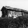 Kvinne i folkedrakt sittende utenfor våningshus, ukjent sted. Stakittgjerde. Huset har skifertak. Slipestein står opp mot husveggen. 
Serie tatt av Robert Collett (1842-1913), amatørfotograf og professor i zoologi.